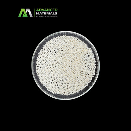 Top-down view of a clear petri dish filled with small white and black spherical particles, centered on a black background with the Advanced Materials by Vivara Scientific logo in the upper left.